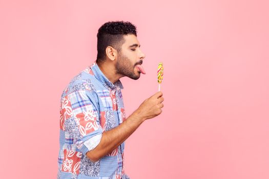 Side view of bearded handsome man wearing blue casual shirt standing tastes lollipop, dreaming licking round sweet sugary rainbow candy.