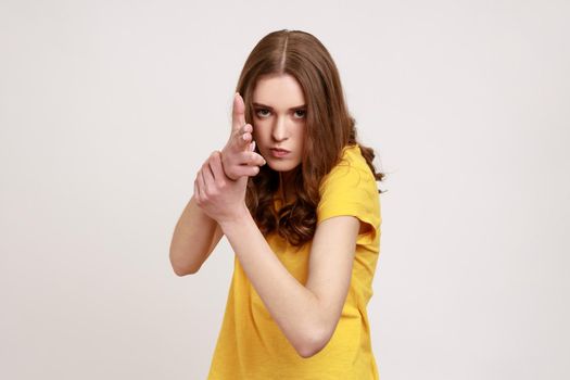 I'll kill you. Portrait of angry teenager girl in yellow T-shirt pointing finger guns to camera, threatening to shoot, hands imitating weapon.