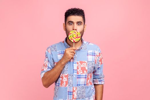 Young adult bearded man wearing blue casual shirt covering his astonished face with round sweet sugary rainbow candy, looking at camera with big eyes.