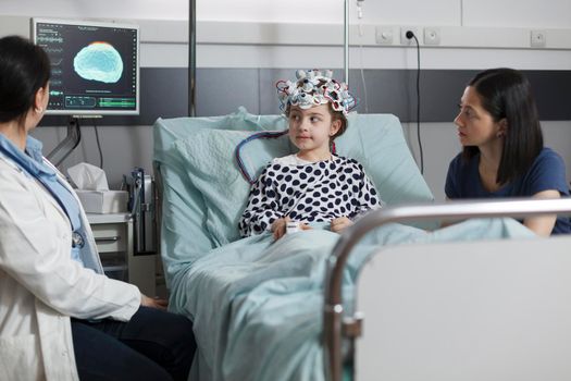 Hospitalized sick girl wearing EEG headset resting in patient bed while doctor analyzing brain condition