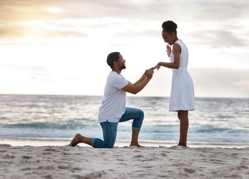 Boyfriend asking his girlfriend to marry him while standing on the beach together. African american man proposing to his girlfriend by the seashore. Young happy couple getting engaged on holiday
