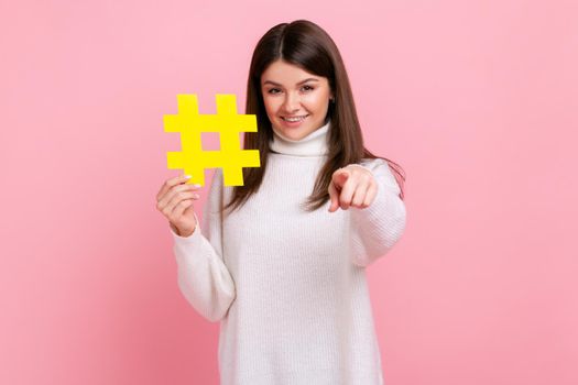 Portrait of happy brunette woman with toothy smile pointing at camera, holding yellow hash symbol.