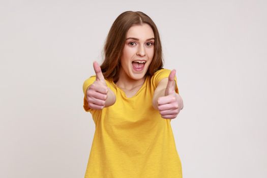 Like. Portrait of enthusiastic woman of young age with brown hair in yellow T-shirt showing thumbs up and yelling excitedly, enjoying excellent result.