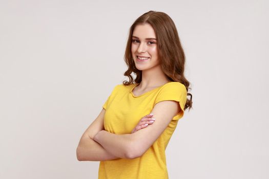 Portrait of positive young woman with brown wavy hair in yellow T-shirt standing with crossed arms, looking at camera with kindness and charming smile.