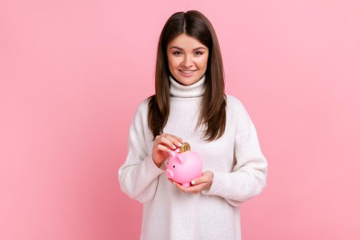 Smiling positive satisfied woman looking at camera, putting coin into piggybank, savings of money.