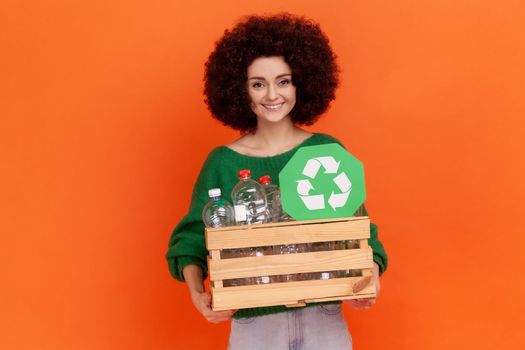 Satisfied woman with Afro hairstyle wearing green casual style sweater with box of empty plastic bottle and green sign, recycle, ecology concept.