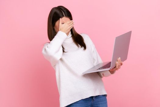 Portrait of brunette female holding laptop computer, covering eyes with palm, sees disgust content.