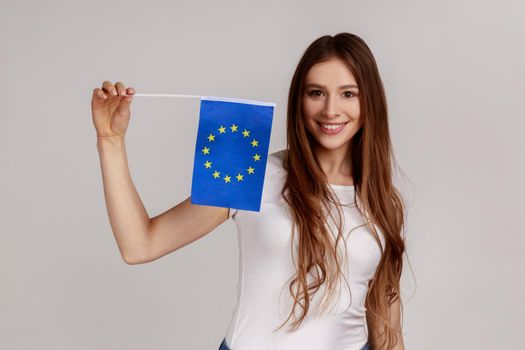 Patriotic woman with dark hair holds eu flag in hand looking at camera with toothy smile, patriotism