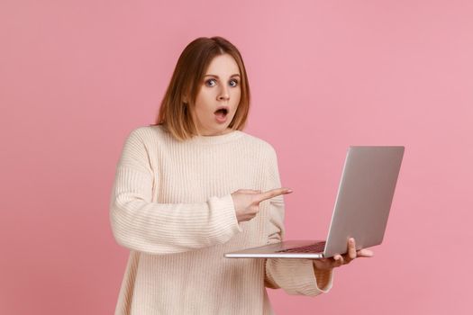 Shocked young adult blond woman working on laptop compute, looking at camera and pointing at display