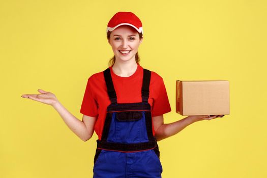 Delivery woman holding parcel and presenting copy space on palm, looking at camera with toothy smile