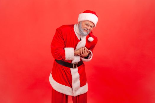 Elderly man with gray beard wearing santa claus costume clutching breast and grimacing from painful cramp, heart attack at young age, cardiac disease.