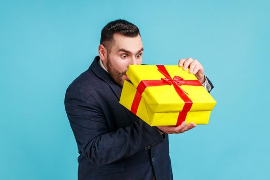 Portrait of curious businessman with present in hands, wearing official style suit, looking inside gift box with shocked expression, unpacking gift.
