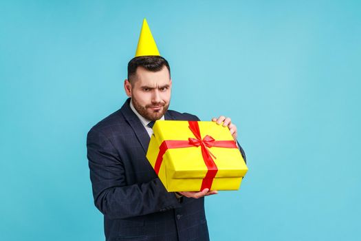 Unhappy man with beard wearing dark suit and party cone opening gift box and looking inside with disappointed sad expression, unwrapping bad present.