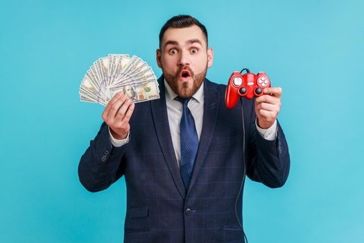 Portrait of extremely shocked businessman wearing official style suit showing to camera red joypad and dollars banknotes, looks surprised.