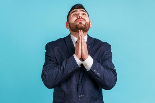 Young adult bearded man wearing official style suit folding hands in prayer, closing eyes and talking to god, asking for help, expressing gratitude.