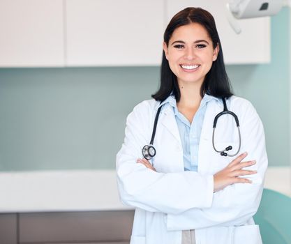 Confident young female dentist standing alone with her arms crossed in a medical office. One caucasian woman in a white coat with stethoscope. Oral hygienist caring for the dental health of patients