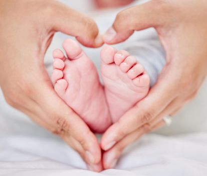 Closeup of hands of a parent forming a heart shape around tiny newborn baby feet. Mother loving her little baby. Small baby resting in its nursery while parent holds its feet.