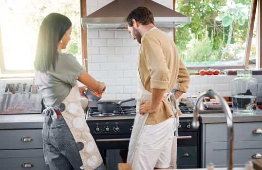 Rear view of young interracial couple cooking a healthy meal together at home. Happy young hispanic wife teaching her husband to cook as throwing vegetables into a pot on the stove