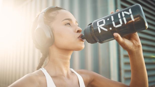 One fit young hispanic woman wearing headphones and taking a rest break to drink water from a bottle while exercising outdoors. Female athlete quenching thirst and cooling down after run and training workout in an urban setting