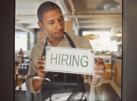 Business owner looking to hire new staff. Young businessman hanging a hiring sign in his shop entrance. Boss working in coffeeshop hiring new employees. Hr employee hanging hiring signage in shop