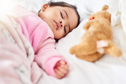Adorable baby girl sleeping peacefully in bed with stuffed animal toy. Baby lying fast asleep on white bed with teddybear and wearing pink clothes and blanket. Mixed race newborn taking a routine nap