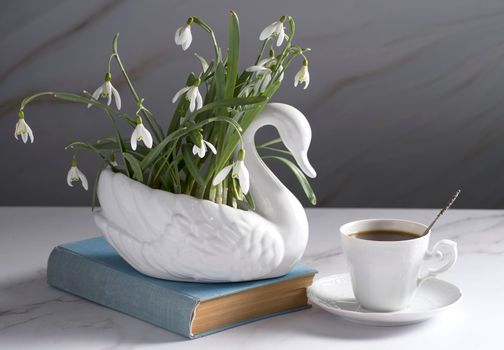 Still life with snowdrops in a ceramic vase like a swan, coffee in a white cup, conceptual morning ritual