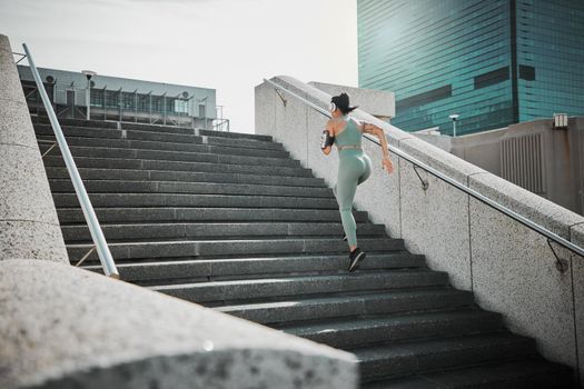 Rear view of a female wearing athlete wearing a phone armband and headphones while running up the steps of a building outside. Young female focused on her speed, body, fitness and cardio health while training