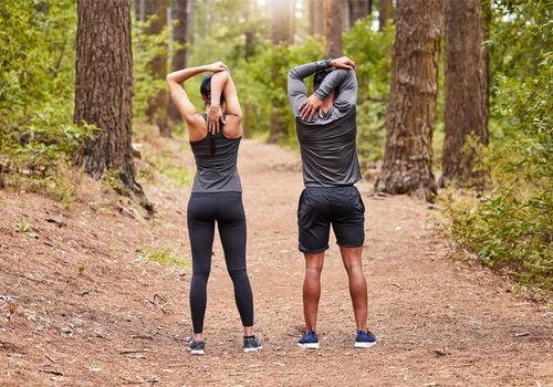 Full length of a young male and female athlete stretching before a run outside in nature from behind. Two fit sportspeople doing warm-up exercises in pine forest on a sunny day