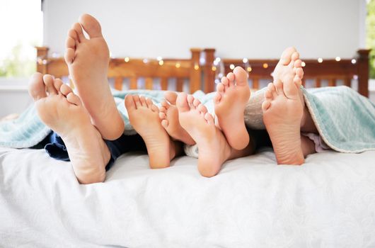 Feet of family lying in bed. Closeup of feet of parents and children in bed. Family relaxing in bed together. Below bare feet of family in bed. Kids resting in bed with their parents