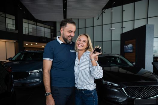 Happy caucasian couple hugging and woman holding new car keys in car showroom.