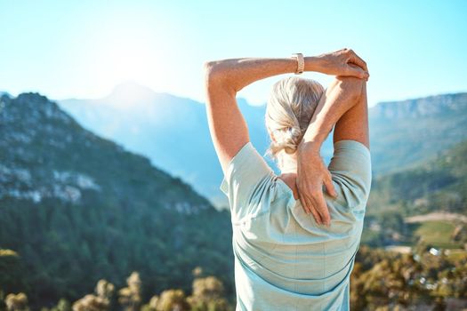 Rear view of a senior woman with grey hair stretching with her hands behind her head while standing outdoors and overlooking a scenic mountain view. Living healthy active lifestyle
