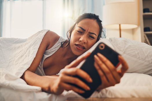 A young mixed race woman looking annoyed while scrolling social media and lying in bed. An attractive Hispanic female using her cellphone and getting bad news while resting in her bedroom