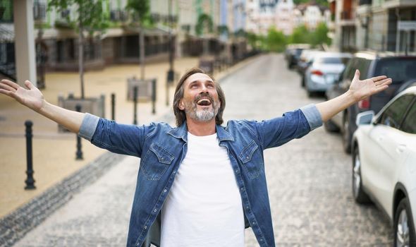Happy senior man thanks God standing outdoors with hands spread wide open wearing jeans shirt with white t-shirt under. Mid Aged man glad that life is beautiful standing in old town street