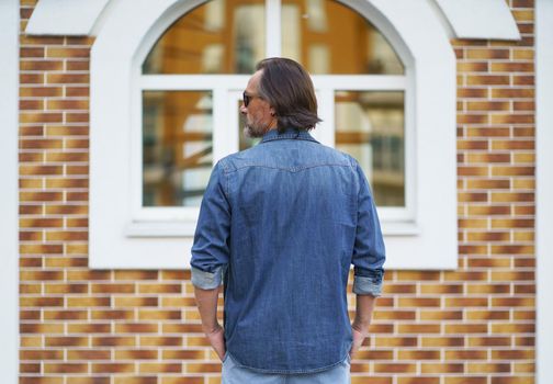 Back view of a man standing alone looking sideways at old town building while traveling in european cities during vacation time wearing denim jeans shirt. Travel concept