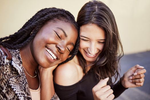 Two beautiful young female friends sitting on sidewalk spending time together in city. Multiethnic best friends looking happy and joyful together
