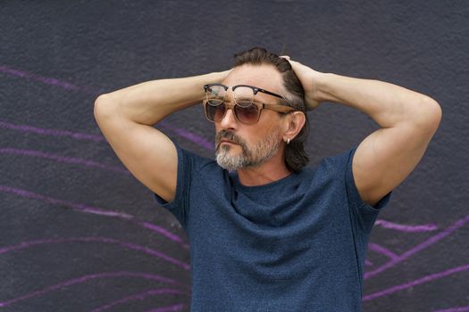 Middle aged man put hands behind head with glasses and sunglasses on his face standing next to dark urban wall wearing dark blue t-shirt. Mature business man or freelancer outdoor