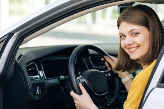 Attractive young business woman looking over her shoulder while driving a car. Driver smiles at camera. Happy millennial woman taxi driver sitting in car holding to steering wheel.