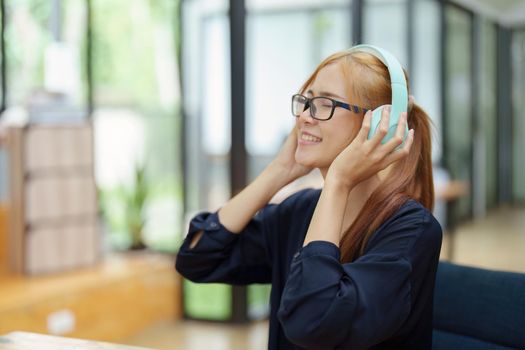 A portrait of a young Asian woman with blonde hair wearing over-ear headphones listening to music to relax while taking a break from boring day activities