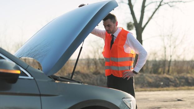 Disappointed man in formal outfit opening bonnet of broke down car to check engine. Sad businessman standing near car opened the hood. Car accident on the road. Emergency stop sign.