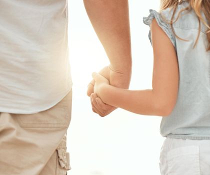 Rear view of a caucasian father holding the hand of his little girl while standing outdoors on a sunny day with a flare in between them. Father and young daughter showing affection and love by holdin