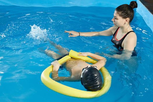 Preschool boy learning to swim in pool with foam noodle with young trainer
