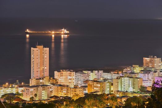 View of skyscraper and city buildings lighting up the dark sky at night, alongside the ocean horizon. Beautiful scenic urban landscape in the evening with a seascape background with a ship at sea.