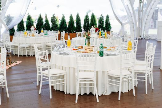 Round dinner tables covered with blue cloth stand in a white wedding pavilion