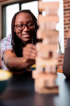 Smiling woman carefully pulling out wood cube from wooden tower structure while sitting at table.