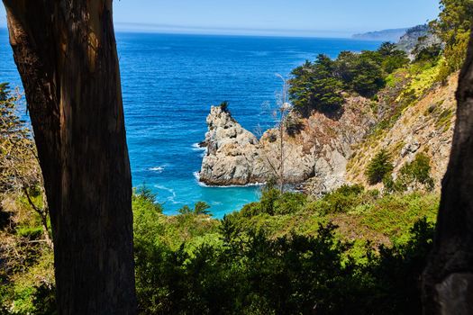 View through trees of ocean coast peninsula with large pillar of rock