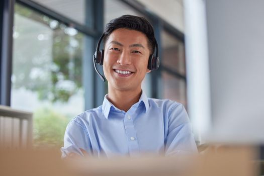 Portrait of a professional asian man smiling and wearing a headset while sitting at his desk in an office job. Happy Asian call centre agent sitting at his desk in an office