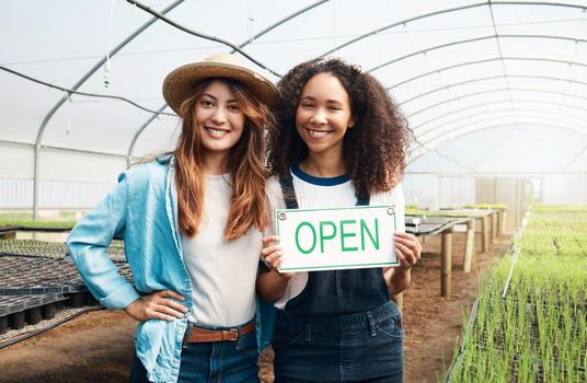 The farm is open for business. Cropped portrait of two attractive young woman holding an open sign while standing in a greenhouse on a farm.