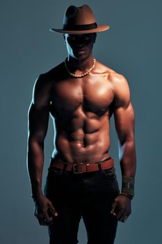 Fashionable african American model posing shirtless against blue studio background with copyspace. Sexy, serious black man with attitude showing bare six pack while wearing hat. Masculine and muscle