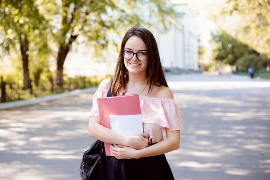 Pretty smiling female student wearing spectacles and casual clothes with books goes to the university to the lecture against background of old conventional university