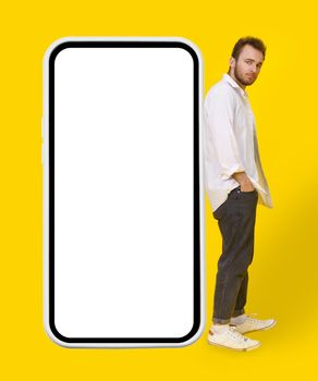 Young man in casual wear and eye glasses leaned on big smartphone posing on yellow background. Stylish bearded smart casual look hipster man with phone. Mobile app advertisement mock up